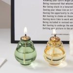 Duas lâmpadas de óleo de vidro, uma verde e uma amarela, com chama acesa, colocadas numa mesa junto a um quadro com texto.