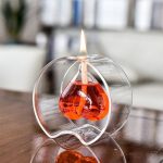 Candeeiro a óleo com forma de coração, com chama acesa, colocado numa superfície de madeira, com fundo de sala de estar.