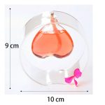 Candeeiro a óleo em forma de coração, com dimensões de 10x9 cm, com vela acesa e laço rosa na base, sobre fundo branco.