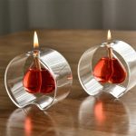 Candeeiro a óleo em forma de coração, com chama acesa, refletindo no vidro transparente, colocado sobre uma superfície de madeira.