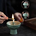 Pessoa acende uma vela de cera numa base de cerâmica verde, com detalhes decorativos, enquanto outra pessoa segura uma tigela de cerâmica com uma vela acesa.