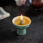 Candeeiro a óleo de cerâmica com chama acesa, colocado numa superfície de madeira escura, com detalhes decorativos na base.