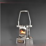 Fogareiro de metal compacto com alça dobrável, aceso com fogo visível na câmara de combustão, sobre fundo escuro.