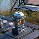 Fogareiro de camping com chaleira de metal, fogo aceso, ao ar livre, com mesa de madeira e cadeiras ao fundo, cenário natural com árvores e céu azul.