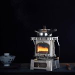 Mini fogão a óleo com tampa metálica, alça e base resistente, usado ao ar livre para cozinhar ou aquecer alimentos.