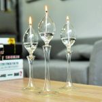 Três candeeiros a óleo de vidro com chamas acesas, colocados sobre uma mesa de madeira, com fundo de sofá e livros.