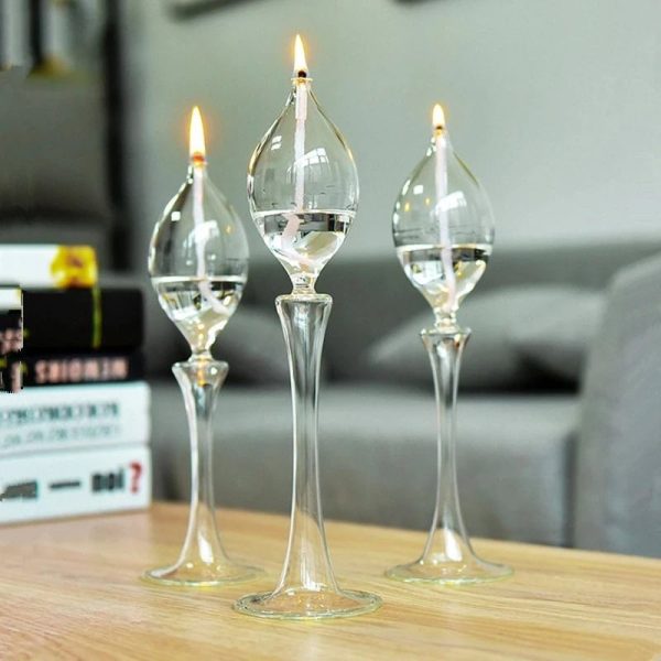 Três candeeiros a óleo de vidro com chamas acesas, colocados sobre uma mesa de madeira, com fundo de sofá e livros.