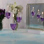 Velas em copos de vidro transparentes com líquido roxo, refletidas num espelho, com flores ao fundo.