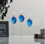 Três candeeiros a óleo de pé com bases de vidro transparente e globos azuis, com velas acesas, colocados numa superfície branca ao lado de uma planta e livros.