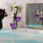 Candeeiro a óleo de pé com chama acesa, refletido num espelho ao lado, com flores ao fundo numa mesa de vidro.