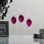 Três candeeiros a óleo de pé com base de vidro transparente e chama acesa, decorando uma mesa moderna com livros e uma planta ao lado.