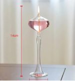 Candeeiro a óleo de pé com base de vidro transparente, corpo alongado e vela acesa na parte superior, altura de 14cm.