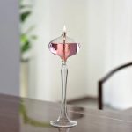 Candeeiro a óleo de pé rosa com chama acesa, colocado numa mesa de madeira, com fundo de cortinas brancas e uma planta verde ao lado.