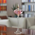 Lâmpada de óleo de pé rosa com chama acesa, colocada sobre uma mesa de madeira, com fundo de sala de estar com livros e sofá.