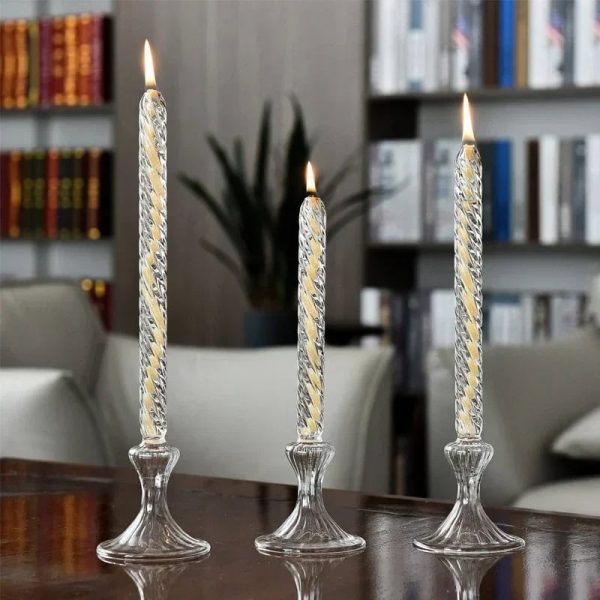 Três velas de cera branca com padrão de espiral acesas em suportes de vidro transparentes em uma mesa de madeira, com fundo de estantes de livros e uma planta.