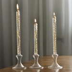 Três velas de vidro decorativas com chama acesa, colocadas em suportes de vidro em diferentes alturas, sobre uma mesa de madeira.