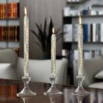 Três velas decorativas de vidro com chama acesa, colocadas numa mesa de madeira, com fundo de estantes de livros e plantas interiores.