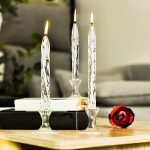 Candeeiro a óleo decorativo com três velas de vidro, uma rosa vermelha ao lado, em ambiente interior com móveis e plantas ao fundo.