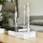Três velas de vidro decorativas com chama acesa, colocadas em suportes de vidro sobre uma mesa de madeira, com livros e decoração ao fundo.
