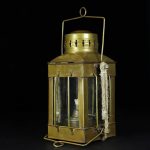 Candeeiro a óleo de latão antigo com vidro e alça de transporte, detalhes de acabamento envelhecido e corda de suporte ao lado.