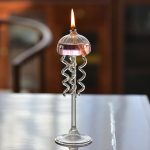 Candeeiro a óleo com design de medusas, com tentáculos de metal e chama acesa, sobre uma mesa de madeira.