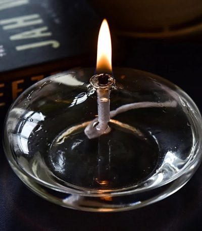 Candeeiro a óleo oval com chama acesa, feito de vidro transparente, com base plana, em uso numa superfície escura, próximo de um livro de ciência.