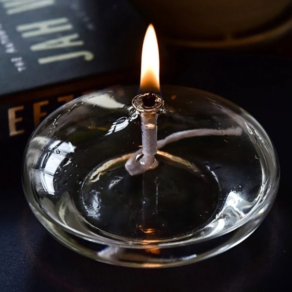 Candeeiro a óleo oval com chama acesa, feito de vidro transparente, com base plana, em uso numa superfície escura, próximo de um livro de ciência.