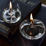 Duas velas de óleo em recipientes de vidro, acesas, com chama brilhante e reflexos na superfície.