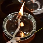 Chama de um candeeiro a óleo oval acesa, com chama longa e brilhante, refletida na superfície de vidro.
