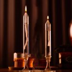 Duas velas de óleo transparentes acesas em suportes de vidro altos, com chama brilhante e detalhes visíveis na cera e no pavio, sobre uma superfície escura.