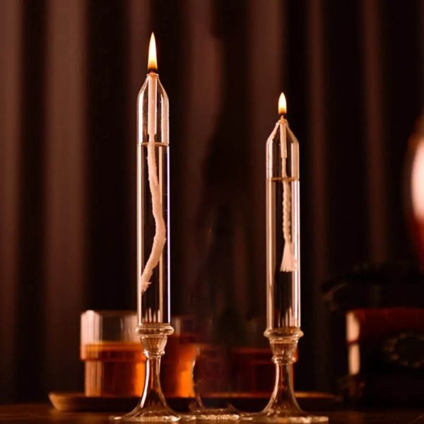Duas velas de óleo transparentes acesas em suportes de vidro altos, com chama brilhante e detalhes visíveis na cera e no pavio, sobre uma superfície escura.