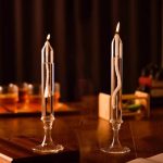 Candeeiro a óleo transparente com chama acesa, colocado numa mesa de madeira, com fundo escuro e objetos de ciência ao fundo.