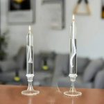 Candeeiro a óleo transparente com chama acesa, visto de frente, com detalhes do pavio e do recipiente de vidro, em ambiente interior com móveis ao fundo.