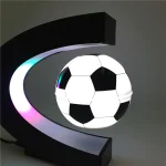 Lâmpada de mesa com design de bola de futebol, iluminação LED colorida, suporte em forma de C preto, ambiente moderno e minimalista.