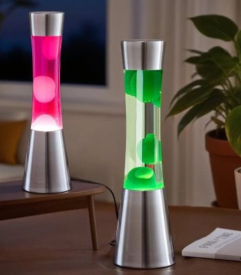 Duas lâmpadas de plasma em vidro com base metálica, uma com luz rosa e outra com luz verde, em ambiente interior com plantas e janela ao fundo.