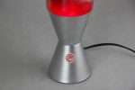 Candeeiro de lava com base metálica prateada, vidro transparente com lava vermelha a subir e descer, ligado a uma fonte de energia elétrica.