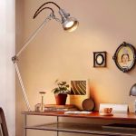 Candeeiro de mesa com braço ajustável e cabeça de foco, iluminando uma secretária com objetos decorativos e livros.