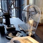 Candeeiro de mesa com design inovador, com uma lâmpada visível dentro de uma estrutura de vidro transparente, colocado numa mesa de sala moderna.