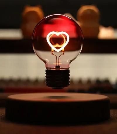 Lâmpada de filamento com coração de néon aceso, refletindo luz quente, com fundo escuro e elementos desfocados ao fundo.