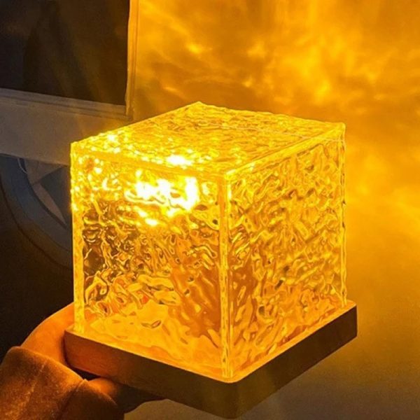 Luminária de mesa em forma de cubo de vidro ambar com luz quente, superfície texturizada, sobre base preta, ambiente acolhedor.