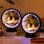 Dois candeeiros de mesa com moldura circular decorada com cristais de sal rosa, refletindo uma sala com flores e objetos decorativos ao fundo.