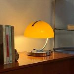 Candeeiro de mesa amarelo com abajur arredondado, base metálica e braço curvo, colocado numa secretária de madeira ao lado de livros e uma pedra decorativa.