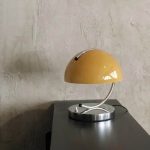 Candeeiro de mesa amarelo com base metálica prateada e braço curvo, iluminação suave, colocado numa superfície escura junto a uma parede de textura clara.