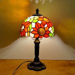 Candeeiro de mesa com base escura e abajur de vitral com padrão de flores vermelhas, amarelas e verdes, iluminado, em ambiente interior.