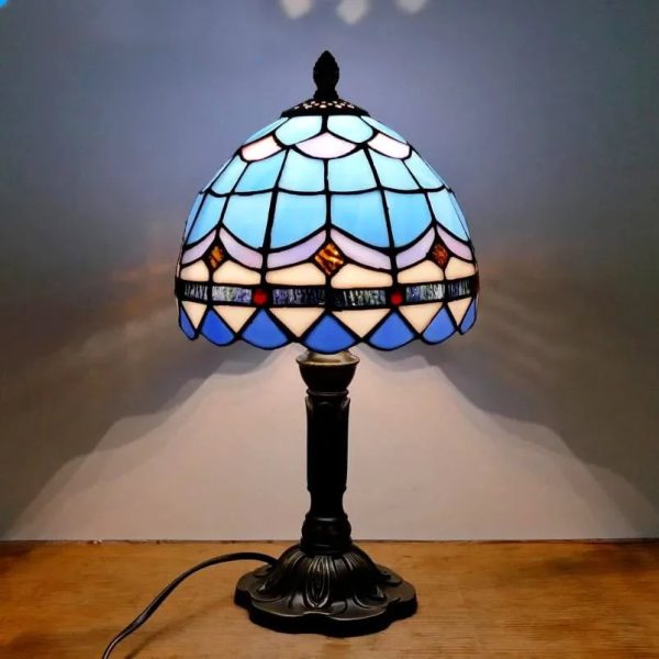 Candeeiro de mesa com base escura e abajur de vitral azul, branco e dourado, iluminado, colocado sobre uma mesa de madeira.