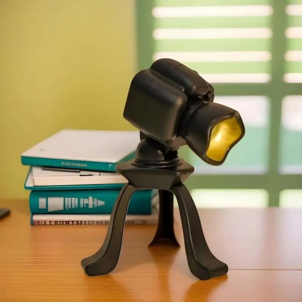 Lâmpada de mesa com corpo de câmara fotográfica, luz amarela, apoiada em tripé preto, ao lado de livros azuis e brancos numa mesa de madeira.