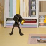 Lâmpada de mesa com design de câmara fotográfica, com corpo preto e lente dourada, colocada numa secretária com livros e materiais de escritório ao fundo.