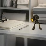 Lâmpada de mesa com design de câmara fotográfica, com luz amarela, colocada numa secretária com livros e materiais de escritório ao fundo.