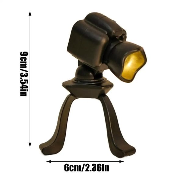 Miniatura de luminária em forma de câmara fotográfica com tripé, iluminação amarela, vista lateral, altura de 9cm, base de 6cm, detalhes em preto e amarelo.