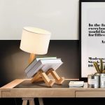 Luminária de mesa com base de madeira em forma de robô, com abajur branco, colocada numa secretária de escritório moderna com quadro e plantas ao fundo.